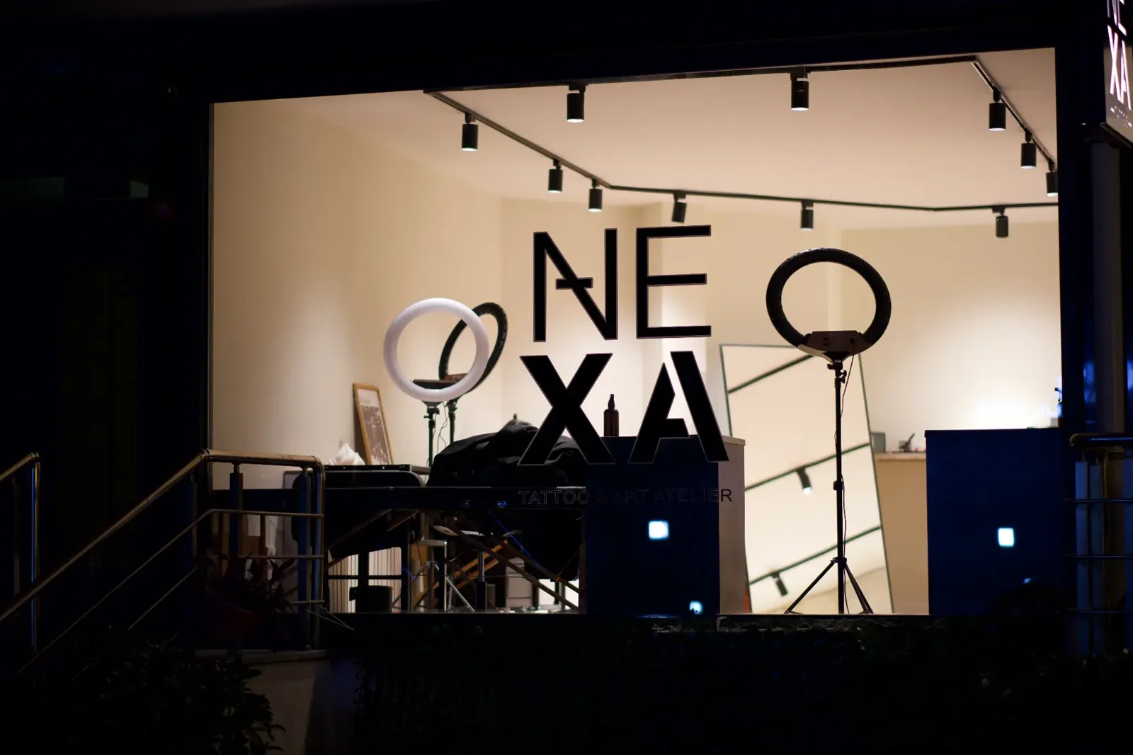 NEXA Atelier gece giriş vitrini — cam bölme ardında NEXA logosu, ring ışıklar ve aydınlatmalı dövme çalışma alanı